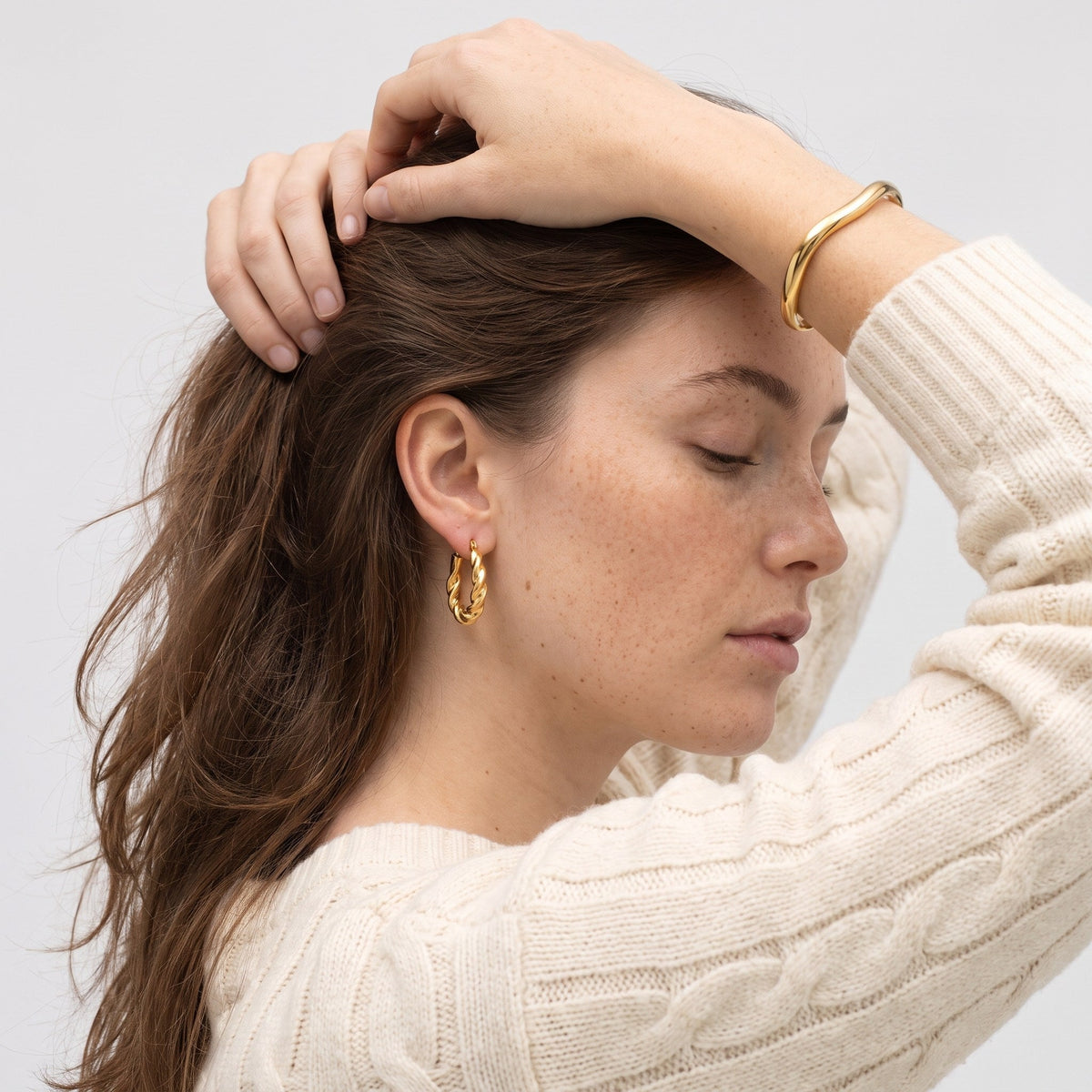 Woman lifting her hair, wearing CYMBRU gold twisted hoop earrings and a gold bangle